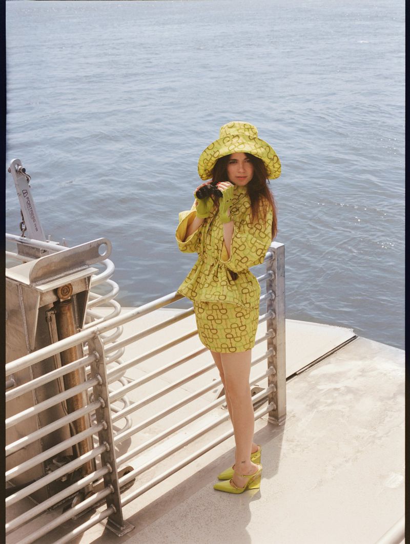A young woman poses on a boat wearing a patterned, lime green mini skirt, jacket, bucket hat, and heels, with water in the background.