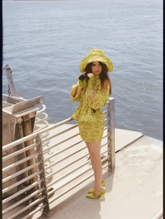 A young woman poses on a boat wearing a patterned, lime green mini skirt, jacket, bucket hat, and heels, with water in the background.