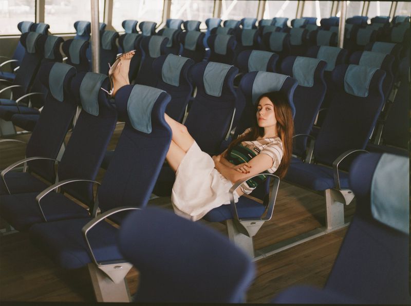 A young woman with long brown hair reclines in a blue passenger seat with armrests on a ferry. Light streams through windows.