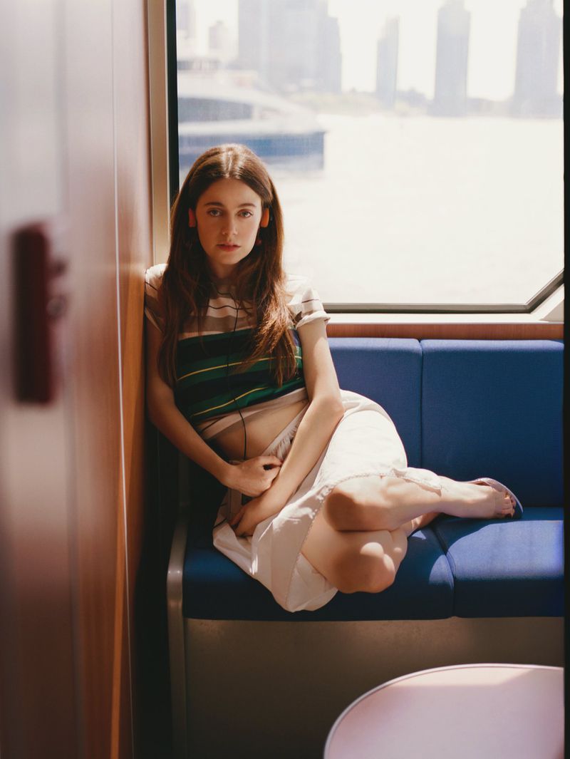 Young woman poses on a blue train seat in front of city buildings. She wears casual clothes and looks at the camera with a neutral expression.
