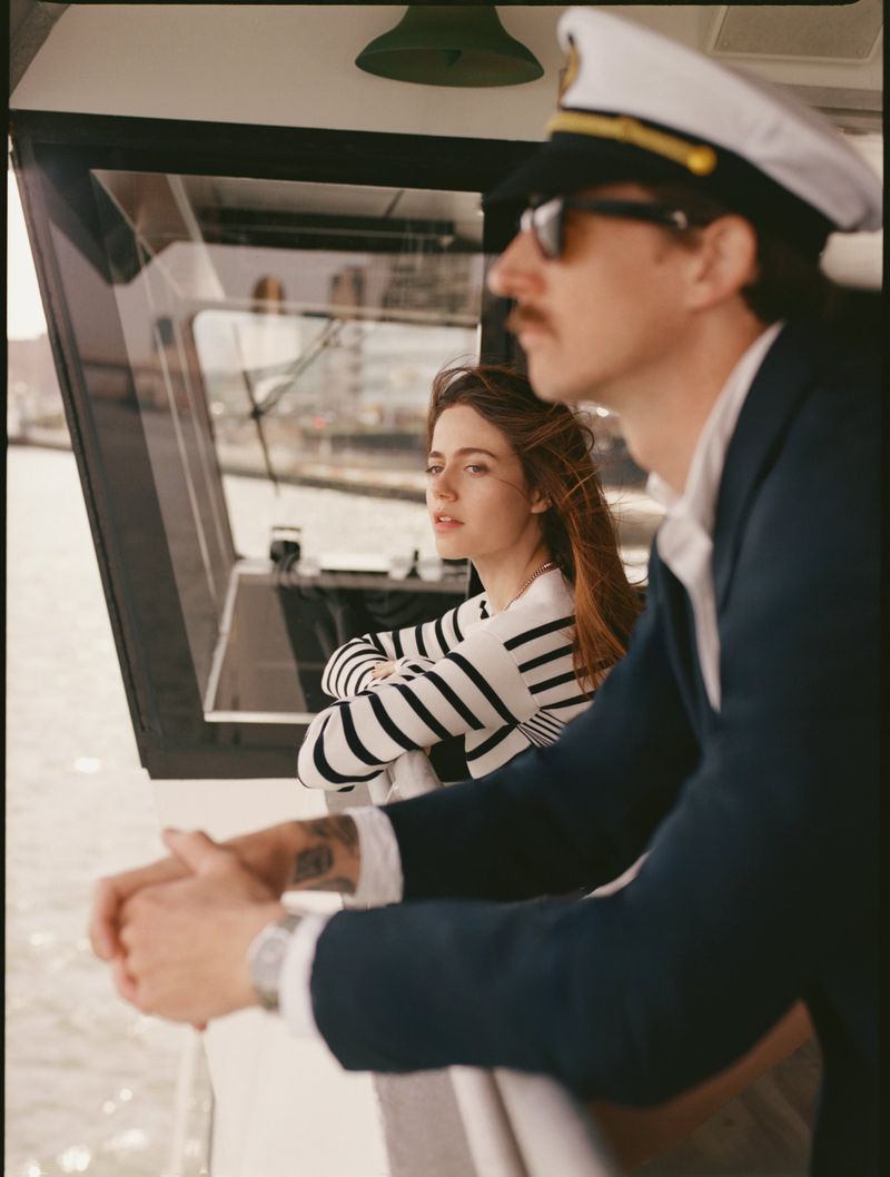 Woman in striped shirt and man in captain's hat and sunglasses on a boat. The pair looks out over a cityscape on a sunny day.