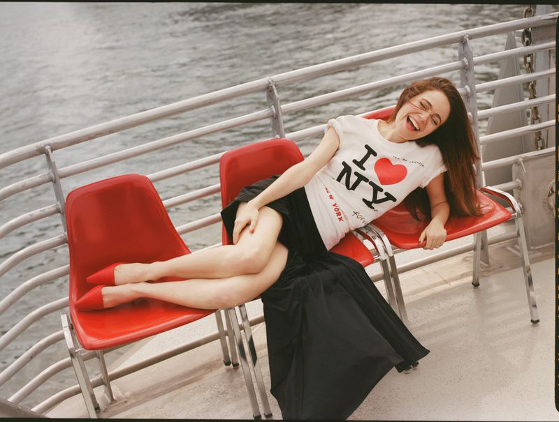 Smiling woman lounges on red chairs on a boat deck wearing an 'I Love NY' t-shirt and long skirt. Water is visible in the background.