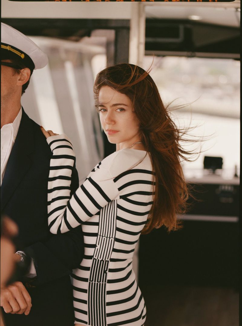 Woman in a striped dress poses with a man wearing a captain's hat on a boat. The woman looks at the camera with a serious expression.