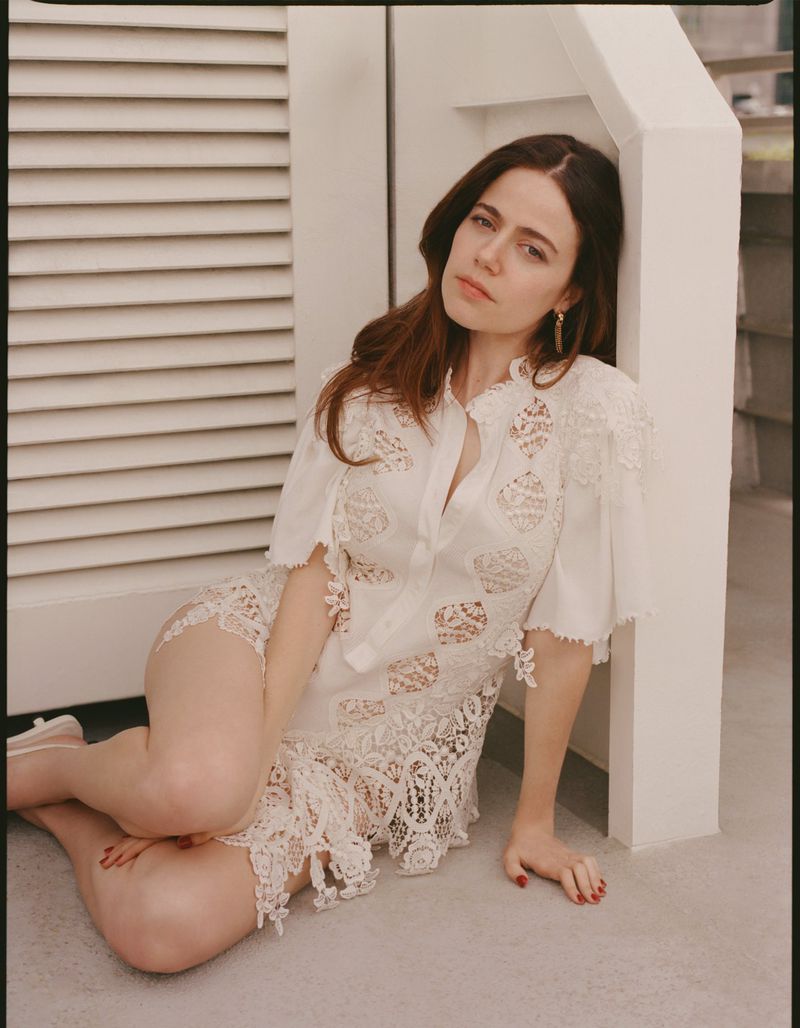 Woman with long brunette hair poses in a white lace dress, leaning against a white wall and shuttered door in an indoor setting.