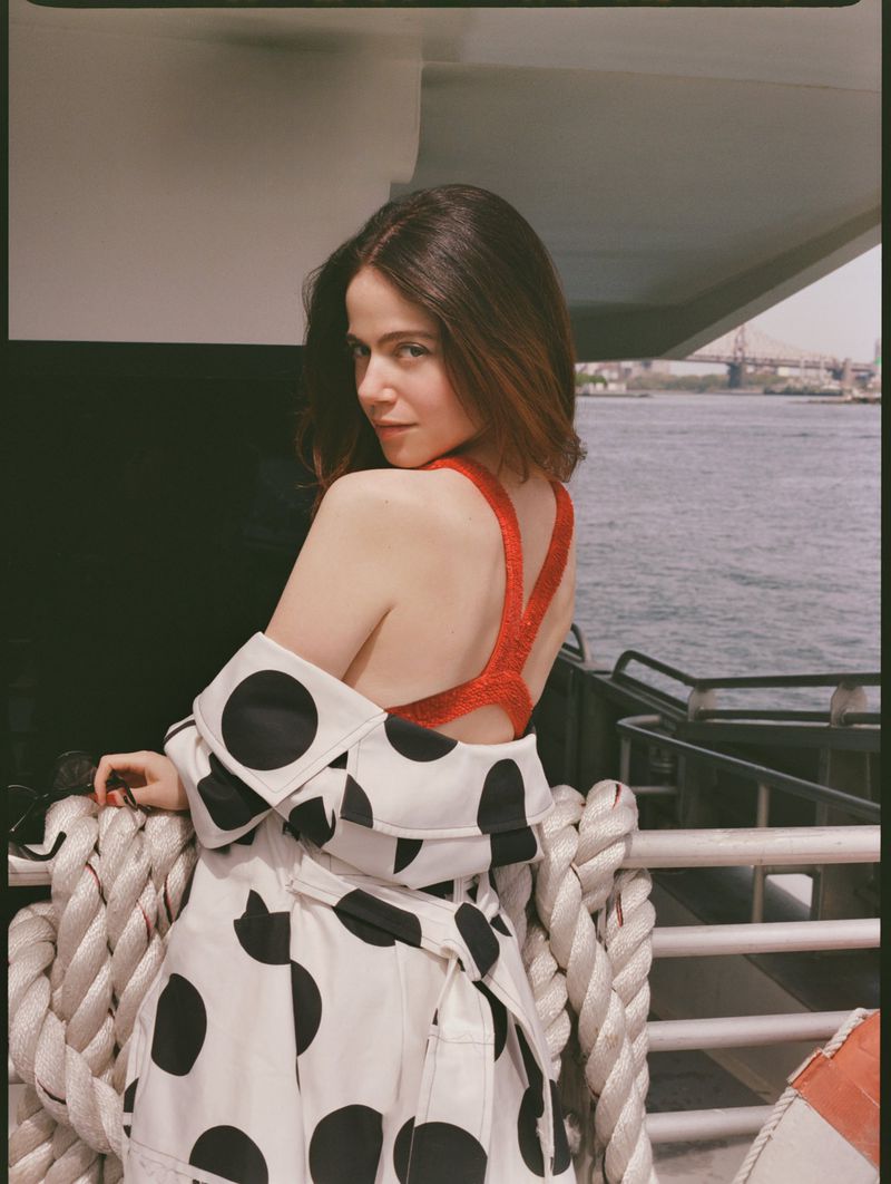 Adult woman in a red top and polka dot coat stands on a boat with brown hair, looking over her shoulder, with city buildings in background.