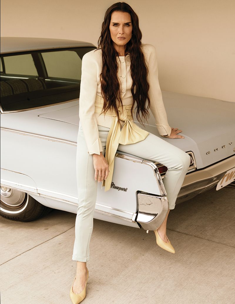 Brooke Shields sitting on the back of a vintage pale blue car wearing a cream blazer with a yellow tied sash and matching trousers.