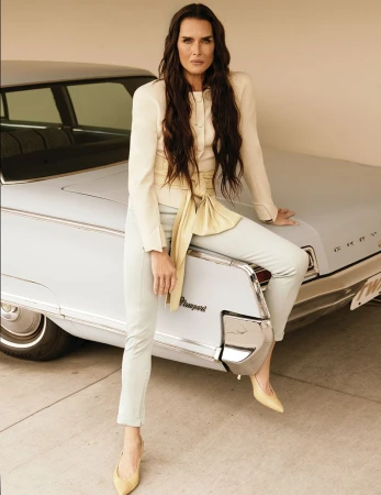Brooke Shields sitting on the back of a vintage pale blue car wearing a cream blazer with a yellow tied sash and matching trousers.