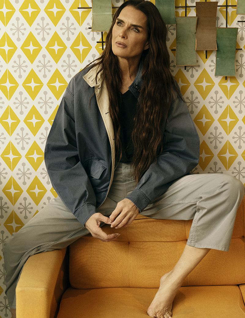 Brooke Shields sitting barefoot in an oversized grey jacket and trousers on an ochre armchair against yellow geometric wallpaper.