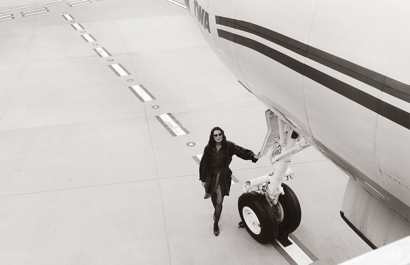 Brooke Shields wearing a dark belted coat and sunglasses while standing beside an airplane landing gear on a runway.