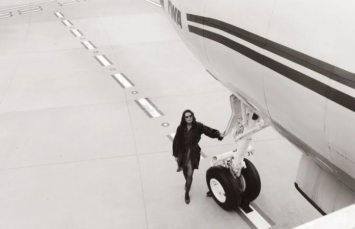 Brooke Shields wearing a dark belted coat and sunglasses while standing beside an airplane landing gear on a runway.