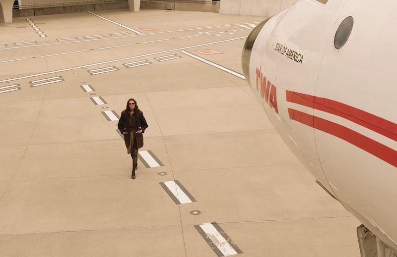 Brooke Shields walking across an airport tarmac in a dark coat and sunglasses beside the nose of a vintage TWA airplane.