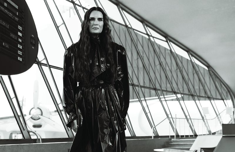 Brooke Shields wearing a glossy black belted trench coat in front of large airport terminal windows overlooking an airplane.