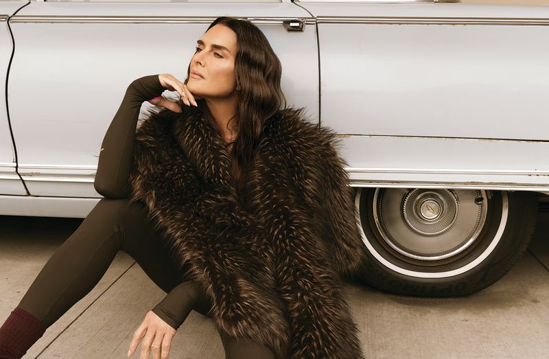 Brooke Shields wearing a dark brown bodysuit and voluminous fur stole while leaning against a vintage car.