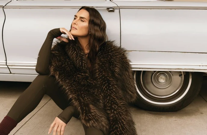 Brooke Shields wearing a dark brown bodysuit and voluminous fur stole while leaning against a vintage car.