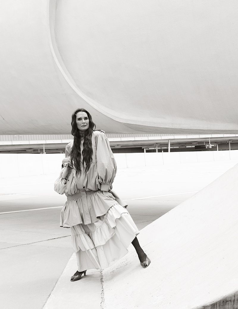 Brooke Shields posing in a voluminous layered tiered dress against curved modern architecture in black and white.