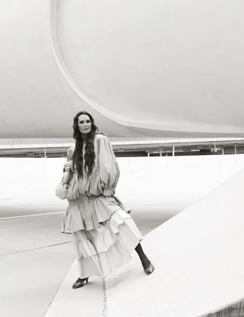 Brooke Shields posing in a voluminous layered tiered dress against curved modern architecture in black and white.