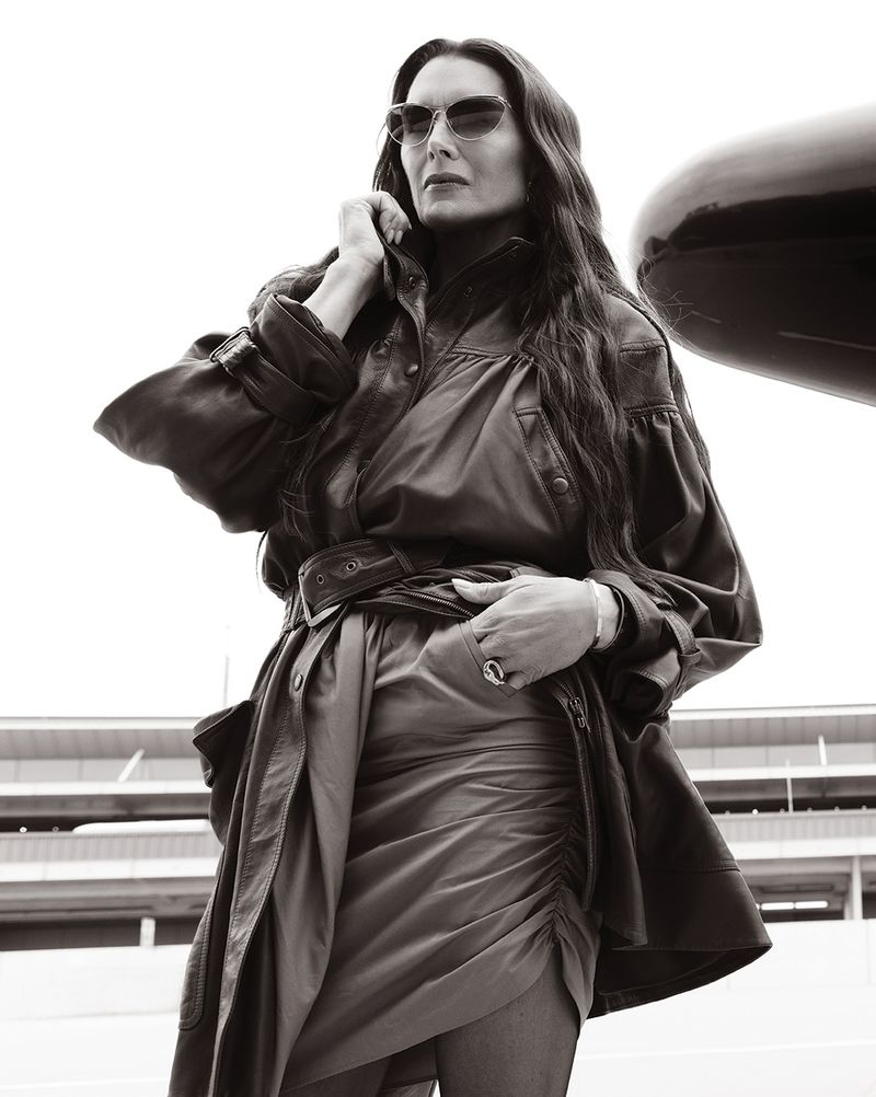 Brooke Shields wearing dark sunglasses, an oversized belted leather jacket, and a ruched skirt in a black-and-white outdoor portrait.
