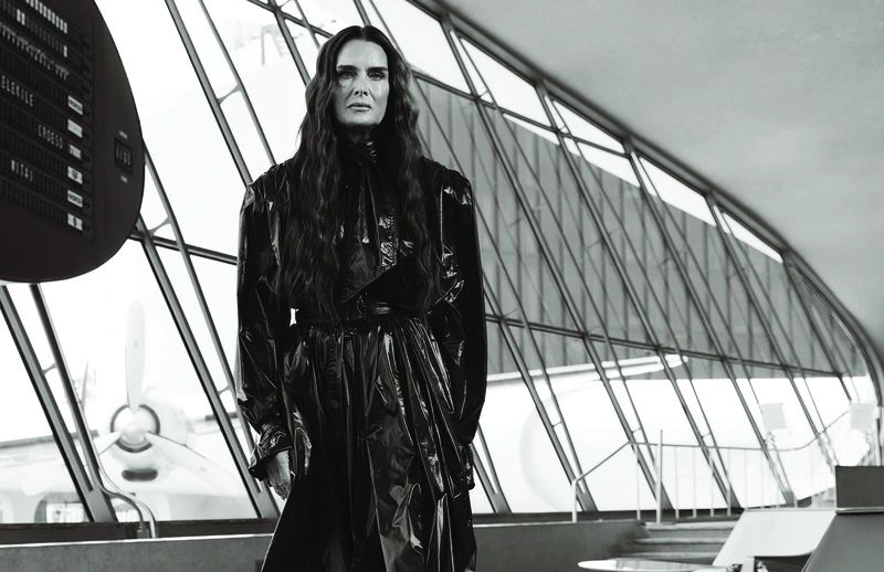 Brooke Shields wearing a glossy black belted trench coat in front of large airport terminal windows overlooking an airplane.
