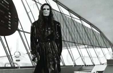 Brooke Shields wearing a glossy black belted trench coat in front of large airport terminal windows overlooking an airplane.