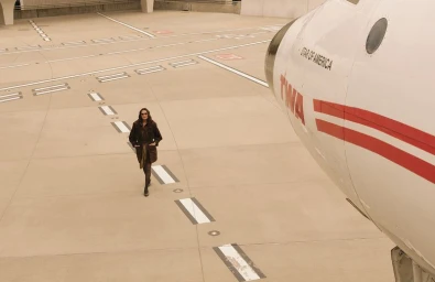 Brooke Shields walking across an airport tarmac in a dark coat and sunglasses beside the nose of a vintage TWA airplane.