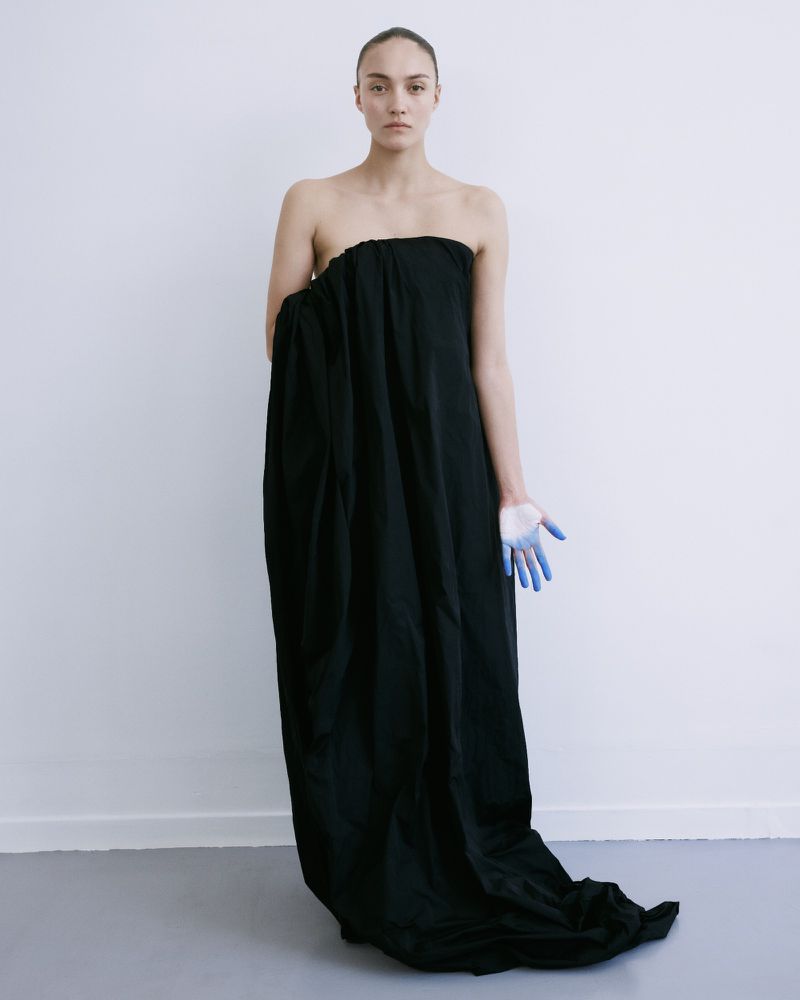 Adult woman in black fabric, one hand with blue paint, against a plain white wall and light gray floor in a minimalist indoor setting.