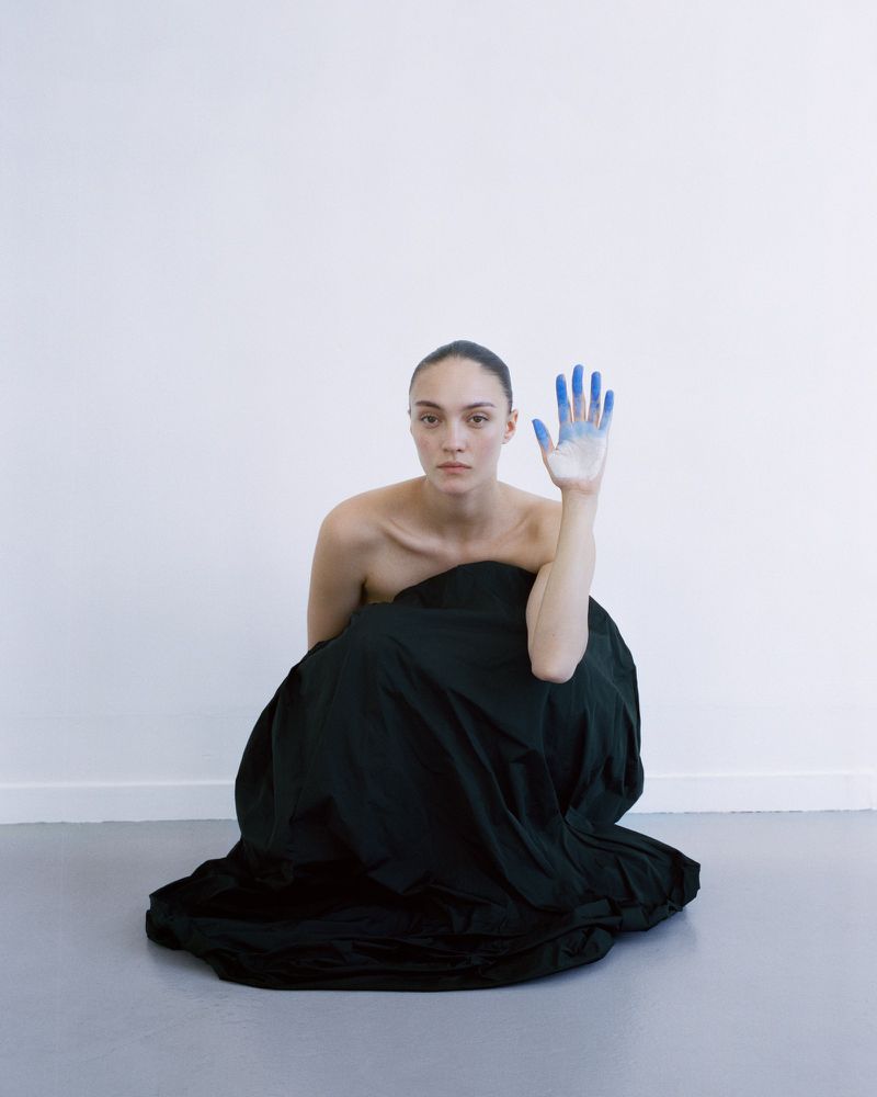 Model wrapped in voluminous black fabric crouching against a white studio wall with blue painted fingers raised.