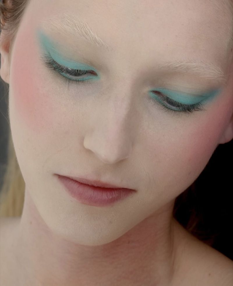 Close-up beauty portrait of a woman looking down, wearing bright turquoise winged eyeshadow, bleached eyebrows, and pink blush.