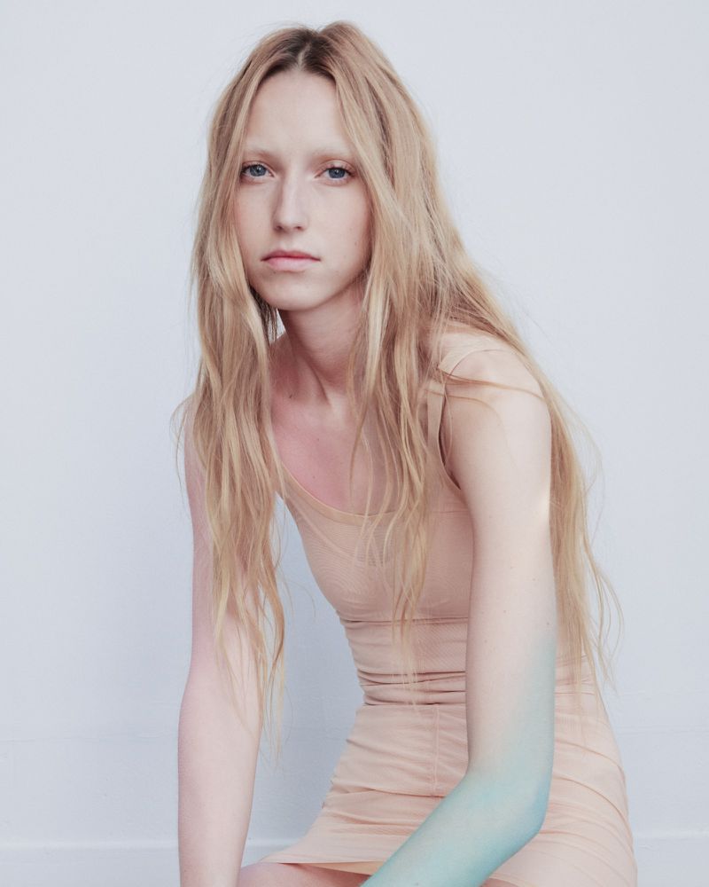 Blonde model in a beige tank dress seated against a light background with subtle blue tone on her arm.