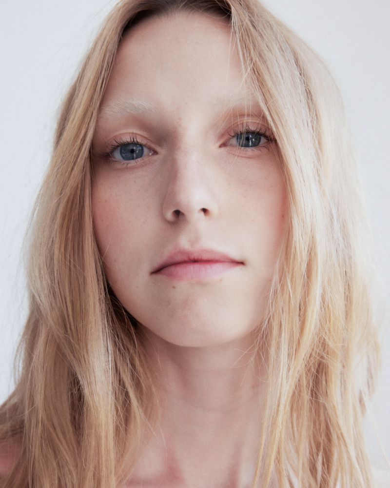 Close-up portrait of a woman with long blonde hair and light blue eyes against a plain white background