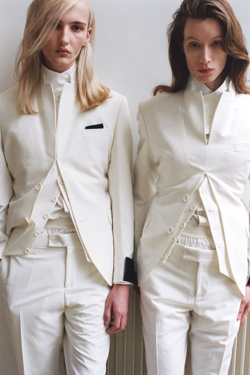 Two models wearing white structured suits standing against a plain light wall in photography by Carlos Folmo