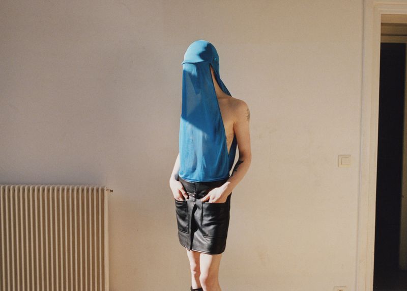 A model standing in a sunlit room with their face covered by a bright blue veil and wearing a black leather mini skirt.