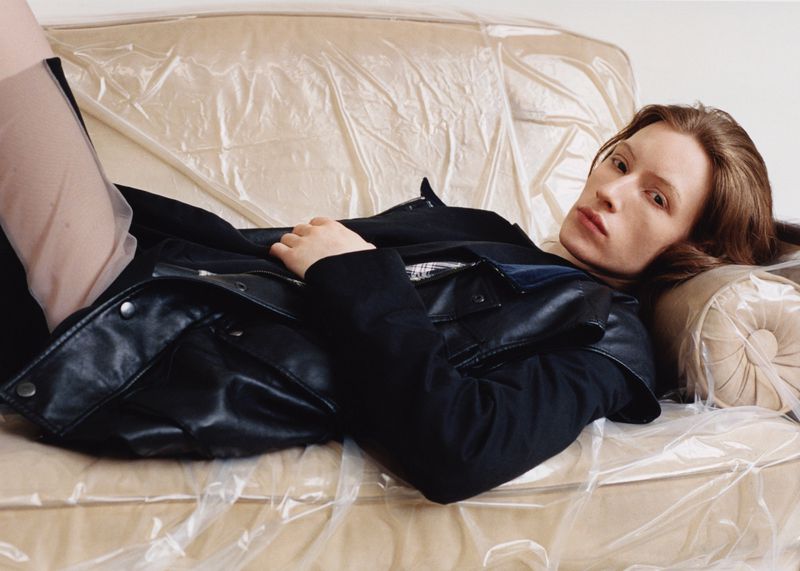 Model wearing a black jacket and leather vest reclining on a beige sofa covered in clear crinkled plastic.