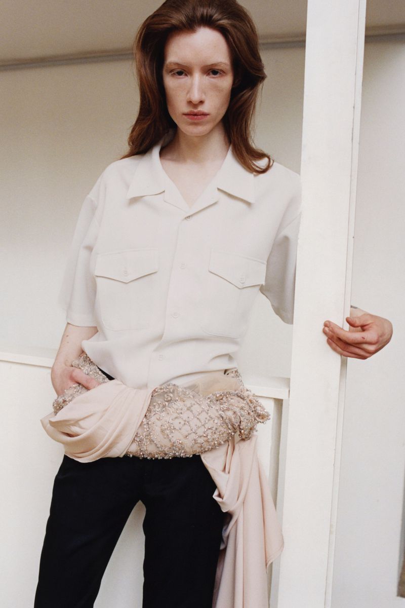 Model with long reddish hair wearing an off-white button shirt and black trousers leaning against a column