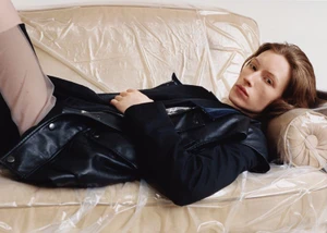 Model wearing a black jacket and leather vest reclining on a beige sofa covered in clear crinkled plastic.