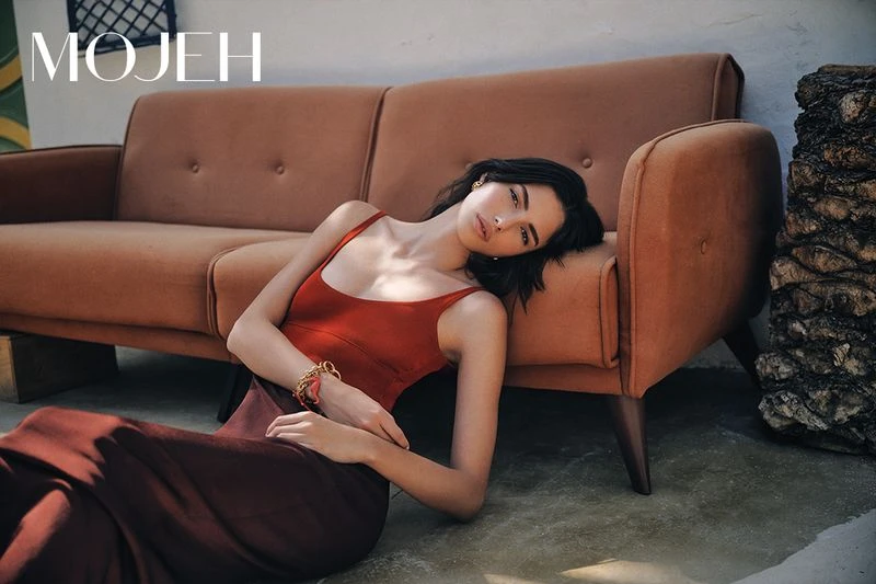 A woman in a rust-colored dress poses on the floor next to a vintage orange couch in a fashion editorial shot with natural lighting.