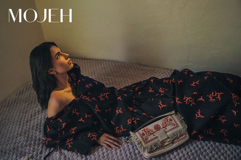 Indoor fashion shot of a woman reclining on a bed wearing a patterned dress, with a handbag and the 'MOJEH' logo visible.