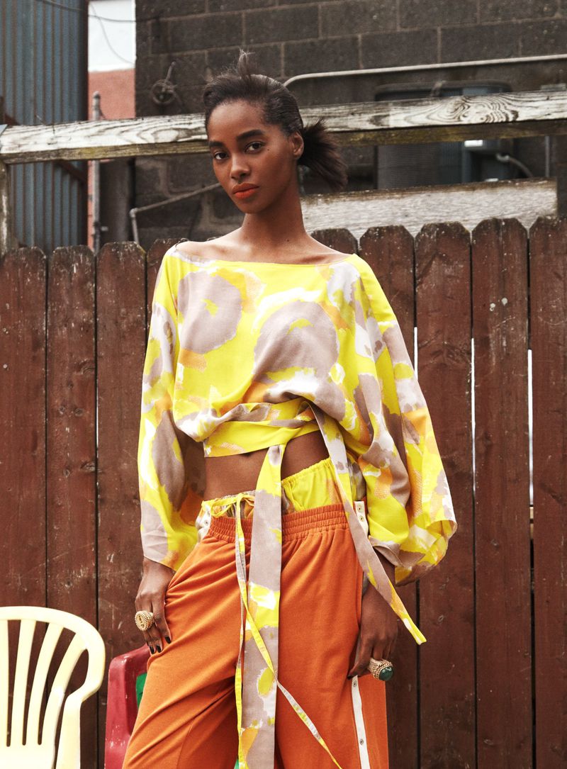 Model Tami Williams wearing a yellow patterned off-the-shoulder top and orange track pants against a wooden fence.