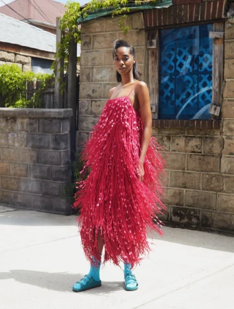 Model Tami Williams wearing a voluminous pink fringe dress with turquoise socks and sandals outdoors in front of a stone building.