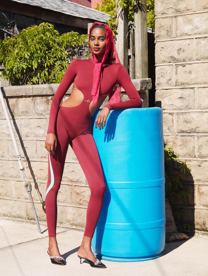 Model Tami Williams in a maroon cutout jumpsuit and pink headscarf leaning on a bright blue plastic barrel outdoors.