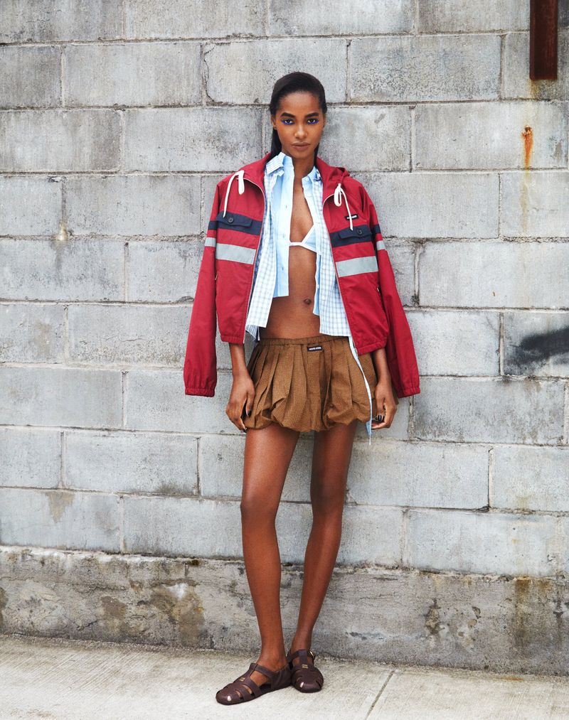 Model Tami Williams posing against a cinder block wall in a red hooded jacket, cropped shirt, and brown pleated mini skirt.
