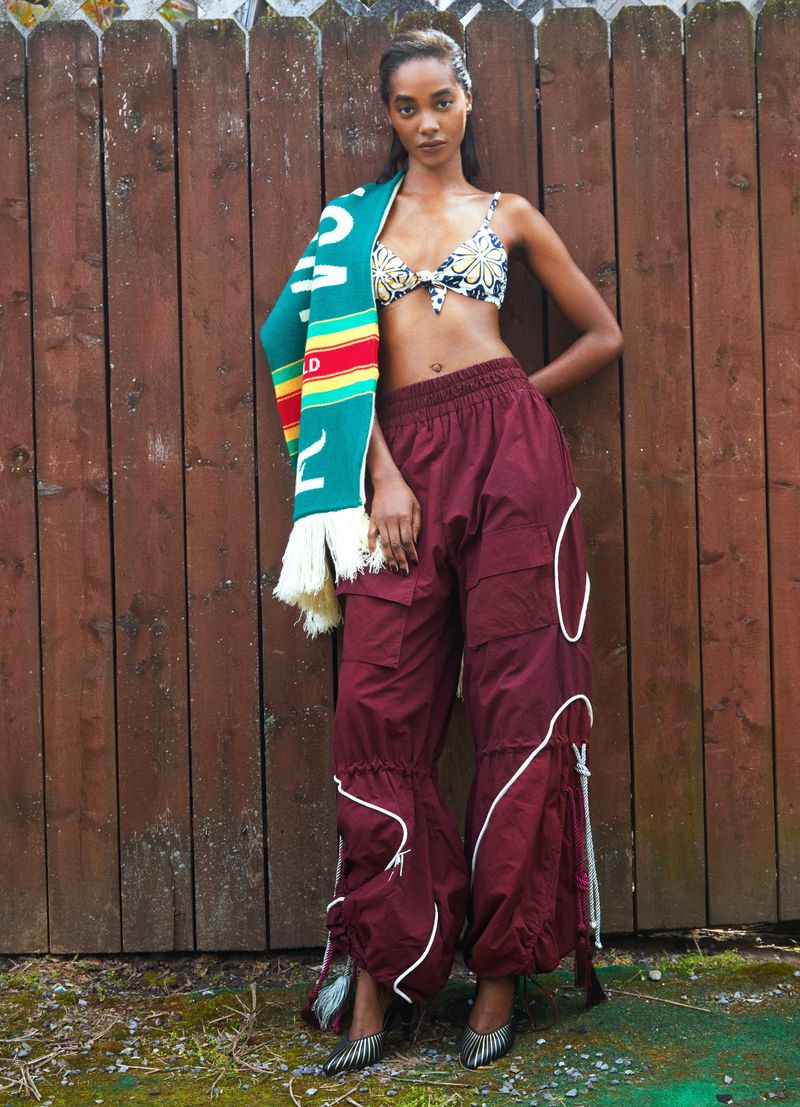 Model Tami Williams poses in burgundy cargo pants, a floral bikini top, and a green scarf against a wooden fence.