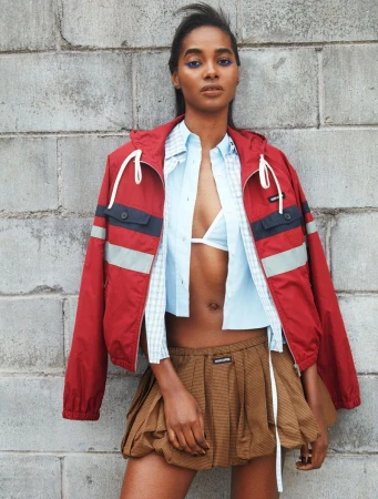 Tami Williams wearing a red hooded windbreaker, open light blue shirt, and pleated brown mini skirt against a concrete block wall.