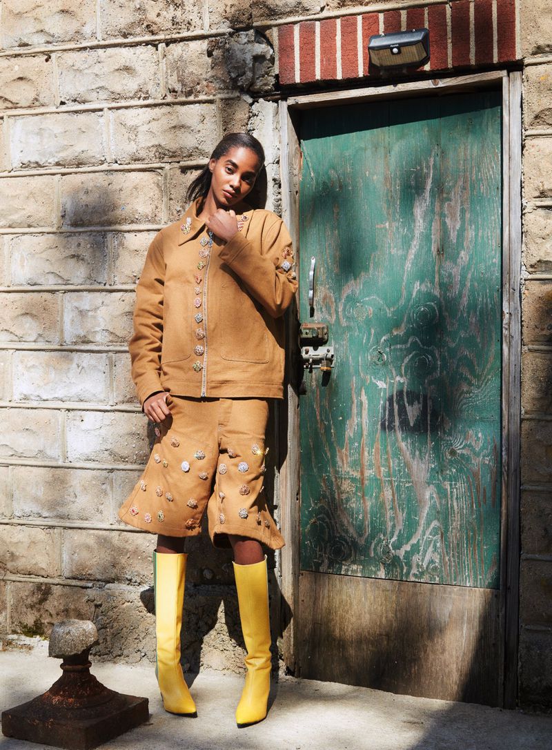 Model Tami Williams wearing tan jacket and yellow boots standing by a green door in Nataal Magazine editorial