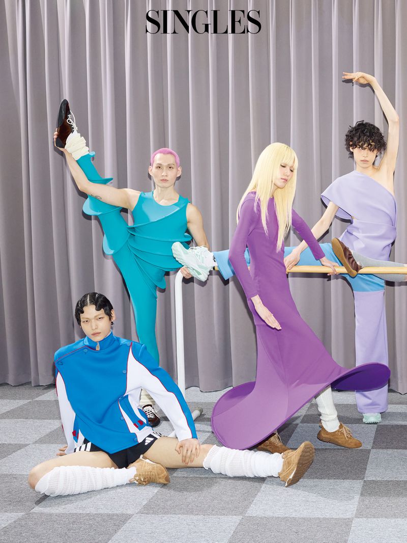 Four fashion models in sculptural purple, teal, and blue outfits posing dynamically around a ballet barre in front of grey curtains.