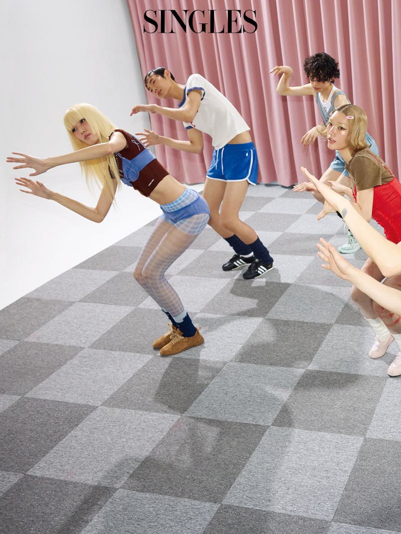 Models pose in dynamic reaching postures on a checkered floor in front of pink pleated curtains for a fashion editorial.