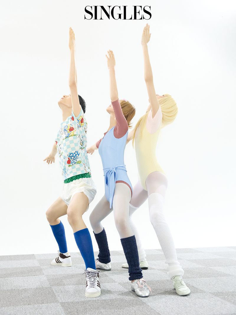 Three models posing in pastel leotards and athletic wear with arms raised on a checkered floor against a white background.