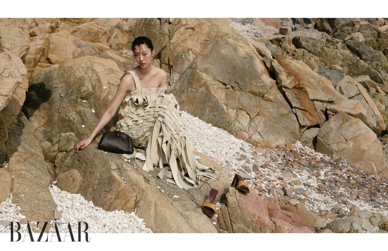 Model Heejung Park reclining on coastal rocks wearing a woven off-white dress with a dark clutch for Harper's Bazaar Korea.