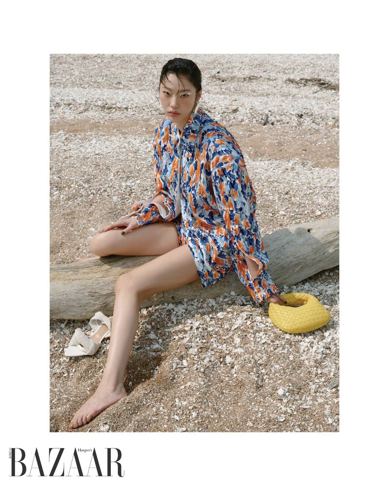 Model seated on a large piece of driftwood on a sandy beach wearing a patterned blue and orange shirt with a yellow handbag.