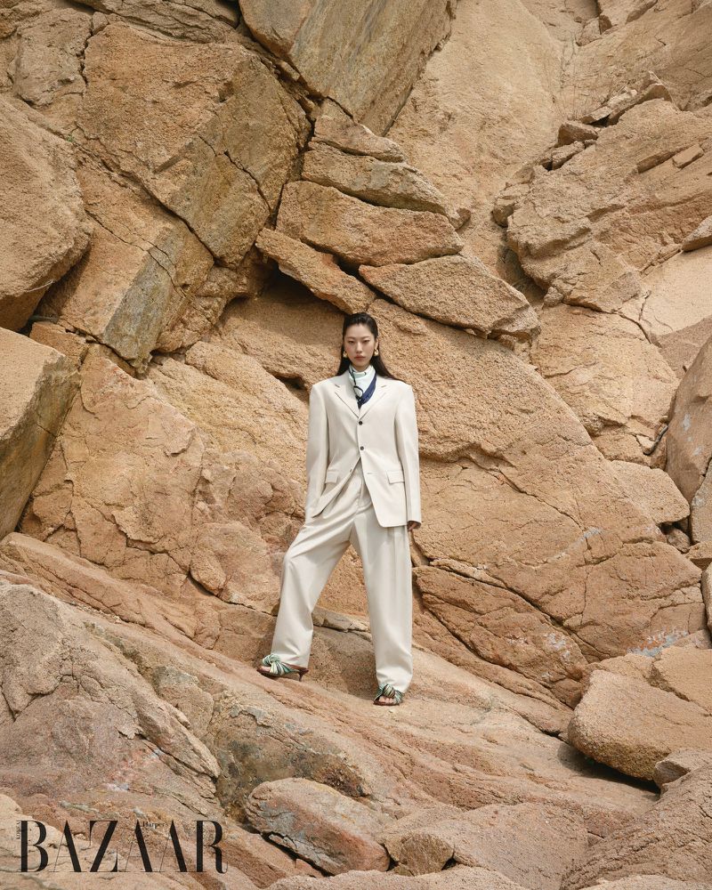 Fashion editorial shot of a young woman in a cream-colored pantsuit posing among large, natural tan rock formations in an outdoor setting.