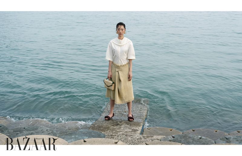Model Heejung Park posing by the ocean in an off-white blouse and beige wrap skirt holding a handbag for Harper's Bazaar Korea.